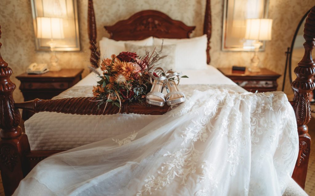 wedding dress, flower and high heels laying on the end of a hotel suite bed